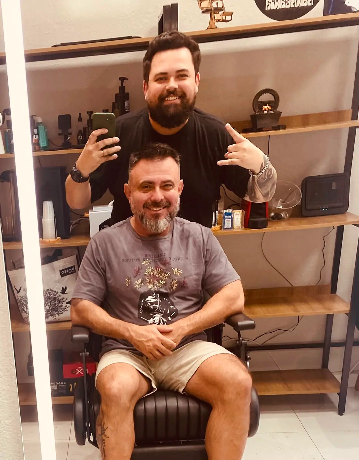 Cliente com barba grisalha aparada após o atendimento, com o barbeiro ao fundo