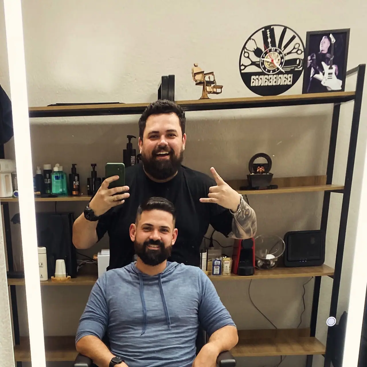 Cliente com corte e barba finalizados, com o barbeiro ao fundo