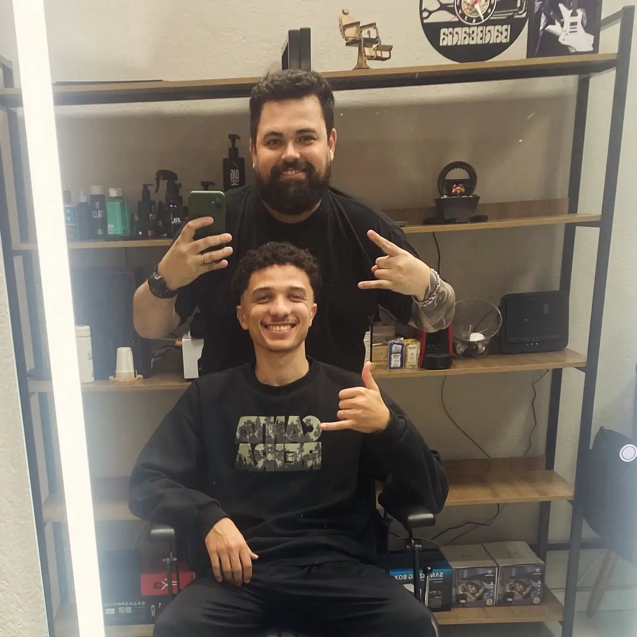 Cliente jovem sorrindo após o corte, com o barbeiro ao fundo